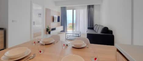 A modern dining area with a wooden table set for four, a sofa in the background, and a balcony view.