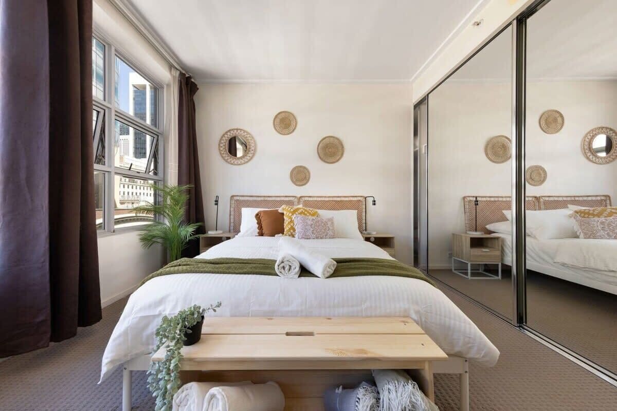 A modern hotel room with a large bed, a bedside table, a bench, and a large window with curtains.