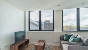 Un salon moderne avec un téléviseur à écran plat posé sur un meuble en bois, un canapé gris agrémenté de coussins verts et une petite table en bois.