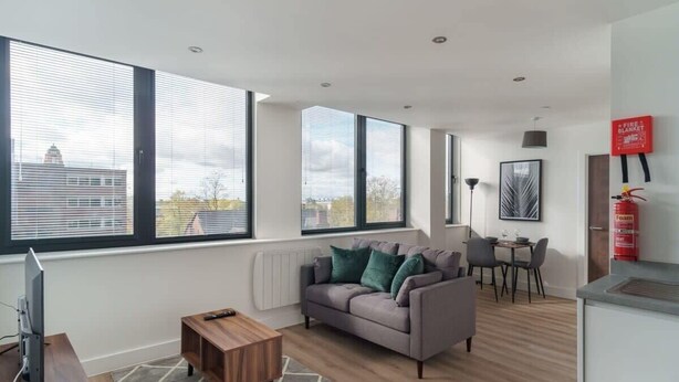 Un salon moderne avec un canapé gris, une table basse en bois et de grandes fenêtres donnant sur un immeuble et des arbres.