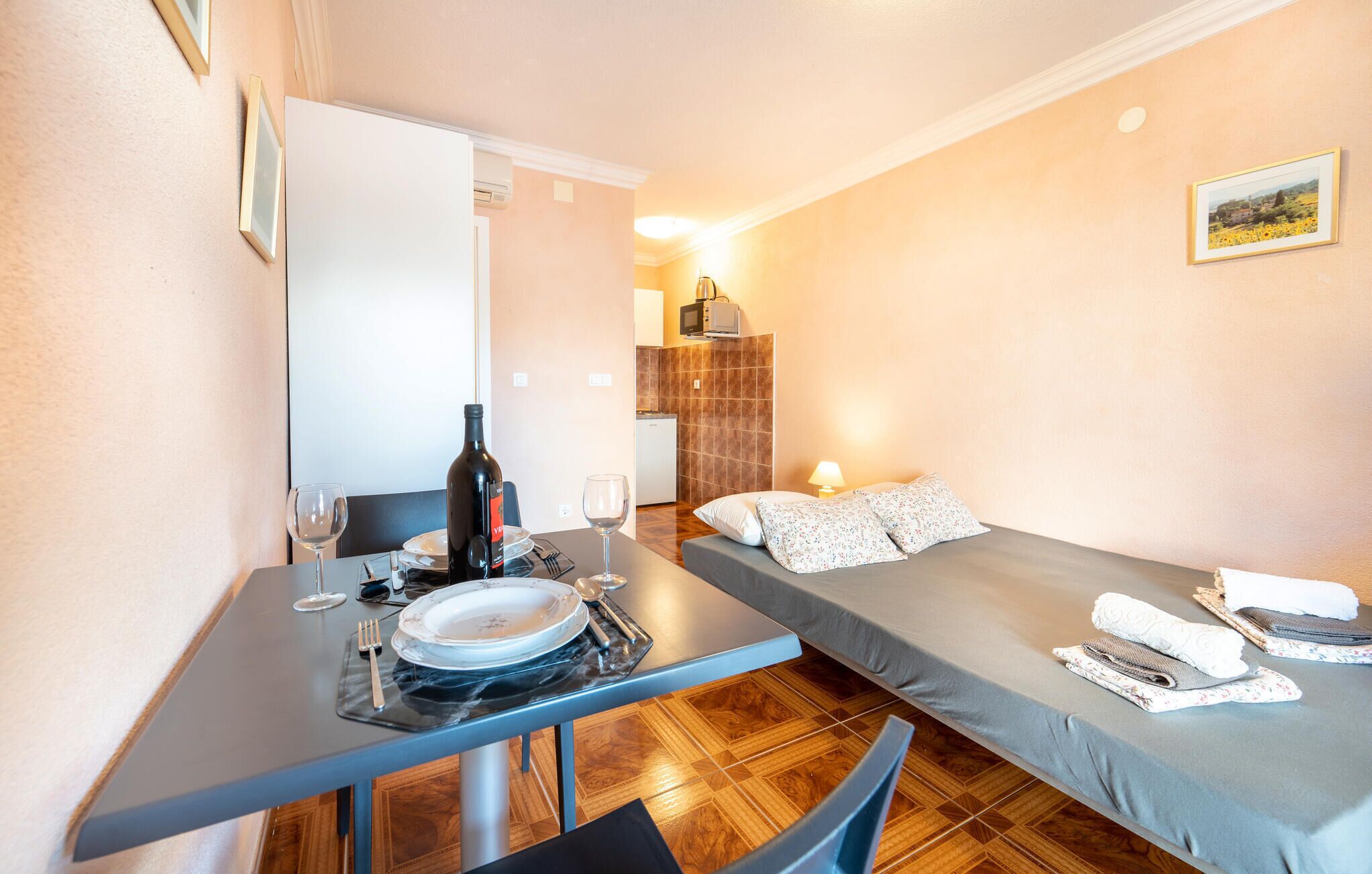 Een kleine kamer met een eettafel gedekt voor twee personen, een bed met kussens, en een kleine kitchenette op de achtergrond.