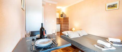 Een kleine kamer met een eettafel gedekt voor twee personen, een bed met kussens, en een kleine kitchenette op de achtergrond.