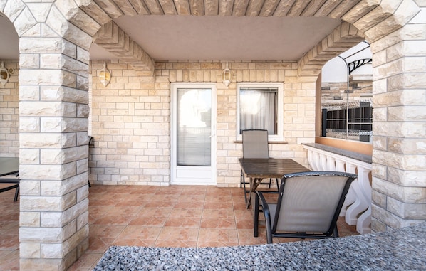 A covered patio area with stone walls, a tiled floor, a table with chairs, and a window with blinds.
