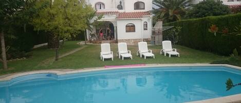 A swimming pool with white lounge chairs, a white building with red-tiled roofs, and a well-maintained lawn.