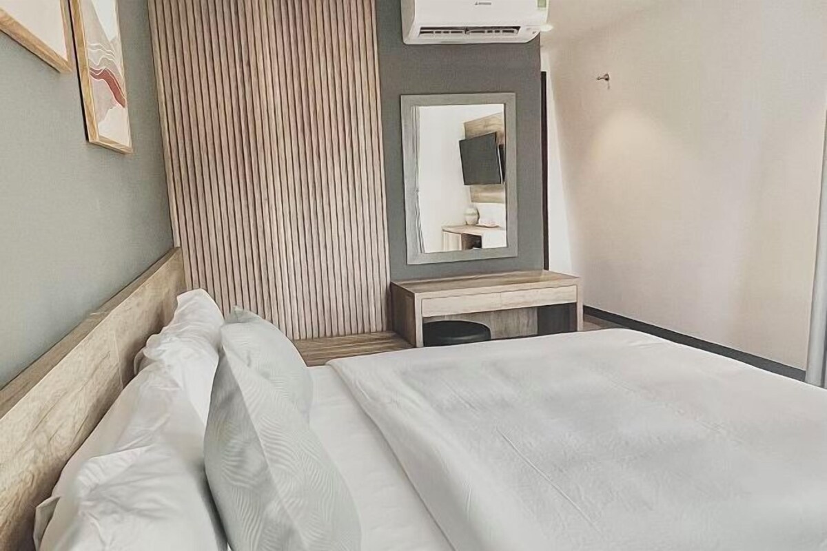 A hotel room with a bed, a bedside table, a mirror, and an air conditioning unit.
