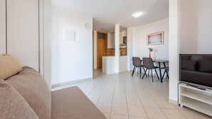 Un salon moderne avec un canapé, un téléviseur, un coin repas avec des chaises et une cuisine à l’arrière-plan.