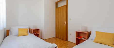 Habitació amb dos llits, cadascun amb matalàs blanc i almoïna groga. Hi ha nits decorades amb llibres i una làmpada a cada costat. Una porta de fusta amb un petit finestral al centre.