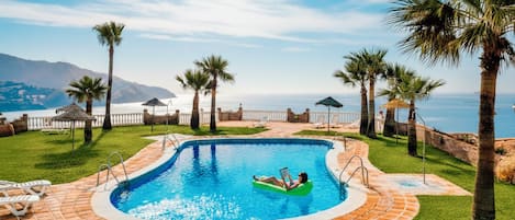 A swimming pool with a person in a green inflatable float, surrounded by palm trees and lounge chairs.