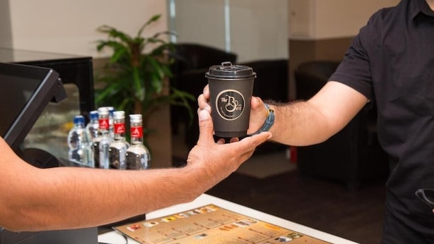 A person holding a coffee cup with a logo, a bar area with bottles, and a plant in the background. - Asis Rise شروق الواحات الراقية للشقق المخدومة (Jeddah)