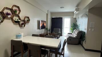 A dining area with a white table and chairs, a sofa, and a massage chair. There are hexagonal shelves on the wall with plants and a painting.