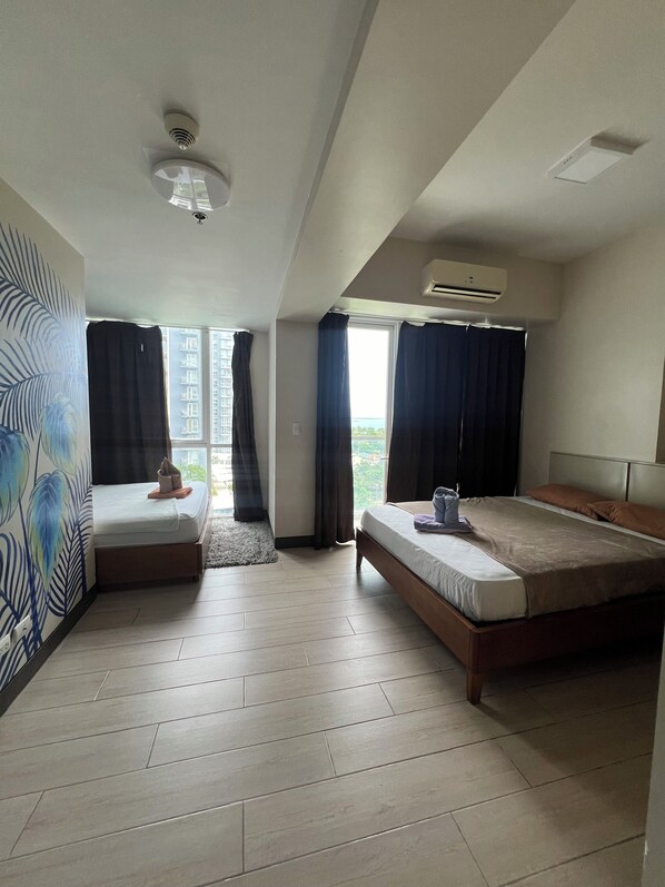 A modern hotel room with a large bed, a smaller bed, and a sofa. There is a window with curtains, an air conditioning unit, and a ceiling fan. - Mactan Newtown Seaview 2 bedroom near airport (Lapu-Lapu)