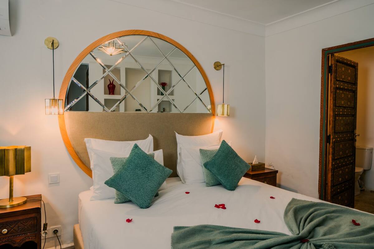 A neatly made bed with green pillows, a large mirror above it, and a bedside lamp.
