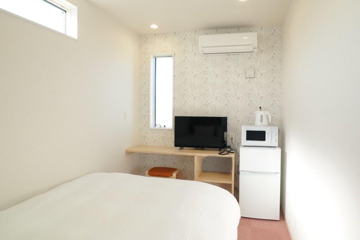 A small, well-lit room with a bed, a desk with a computer, a microwave, and a refrigerator.