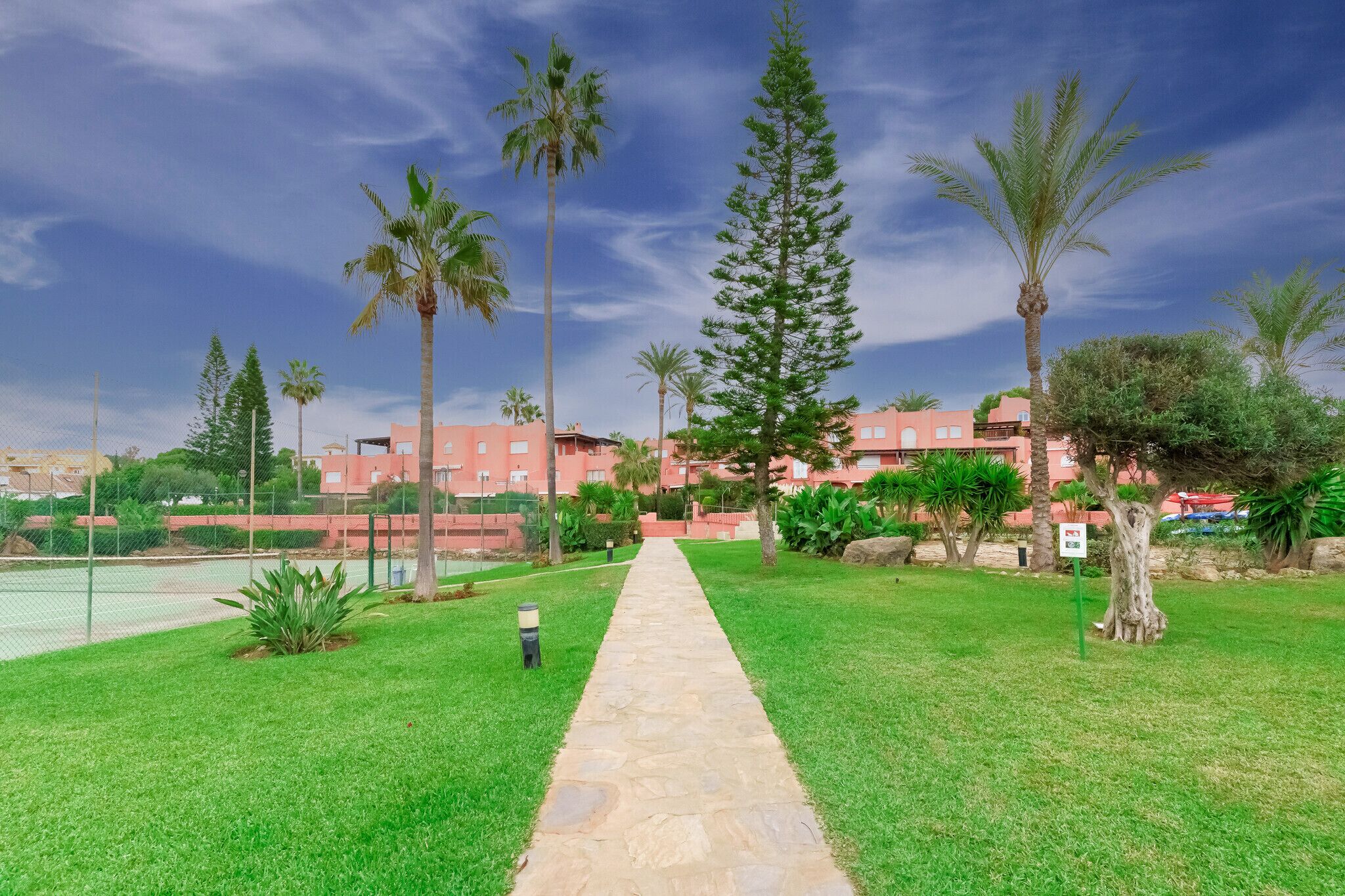 A pathway leading to a building complex with a tennis court visible in the background.