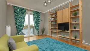 A living room with a green sofa, a blue shag rug, wooden shelves with plants, and a TV. - Comfortable Apartment in Warsaw | Balcony and Parking (Warszawa)