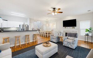 A modern living room with a fireplace, a kitchen with white cabinets, and a dining area with wooden chairs.