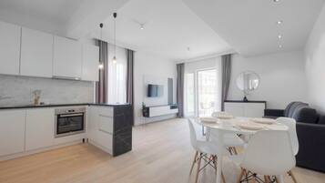 Una cocina moderna con horno empotrado, un comedor con mesa y sillas blancas, y una sala de estar con sofá y televisor.