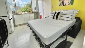 Un dormitorio con cama, ventana con vistas, zona de cocina y pared amarilla con un cuadro.