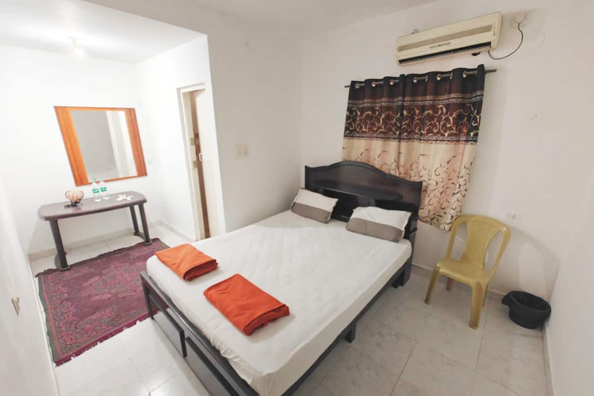 A bedroom with a bed, a bedside table, a mirror, an air conditioner, and a chair.