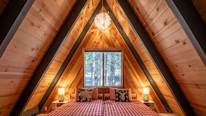 Um quarto de sótão aconchegante com teto de madeira inclinado, uma janela grande com vista para árvores, duas mesas de cabeceira com abajures e roupa de cama xadrez.