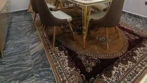 A dining area with a glass table surrounded by chairs, a patterned rug, and a ceiling light fixture.