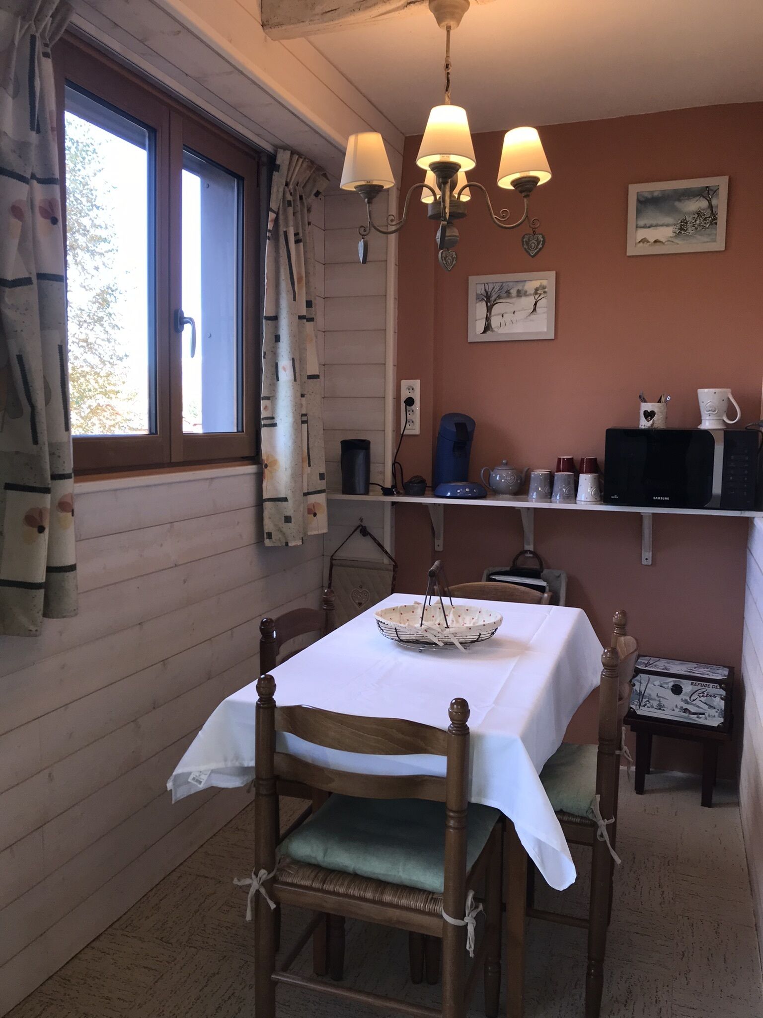 Un coin repas chaleureux avec une table dressée pour deux personnes, un lustre et une fenêtre agrémentée de rideaux.