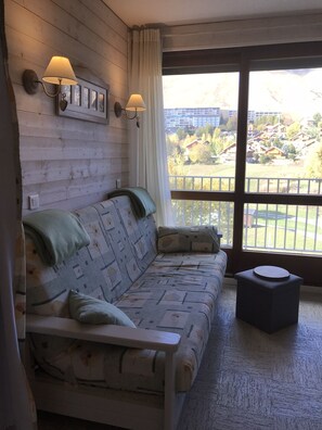 Ein gemütliches Zimmer mit einer Holzwand, einem Sofa mit gemusterten Kissen, einem kleinen Tisch und einem großen Fenster mit Blick auf Häuser und Bäume.