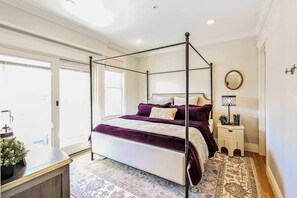 Una cama con dosel con una colcha blanca y morada, una mesita de noche con una lámpara, y un ventanal con vistas al exterior.