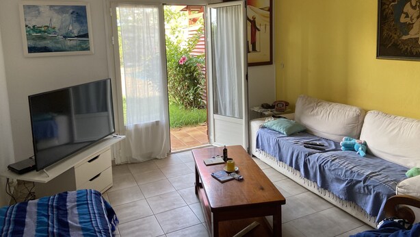 A living room with a TV, sofa, and a wooden table. There is a door leading to an outdoor area with plants. - Location Saisonnière Grand Espace Familial. Plage, Activités Nautiques à 5 mn (Le Moule)