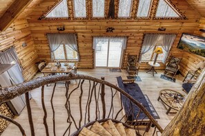 A cabin interior with a spiral staircase, wooden walls, a dining area, and a large window overlooking a snowy landscape.
