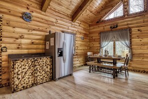 Interior de una cabaña con detalles de madera, un refrigerador de acero inoxidable, una mesa de comedor con sillas y una ventana con cortinas.