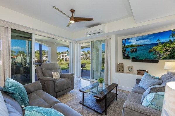 A spacious living room with a large sliding glass door leading to a balcony, a ceiling fan, and a large flat-screen TV mounted on the wall.