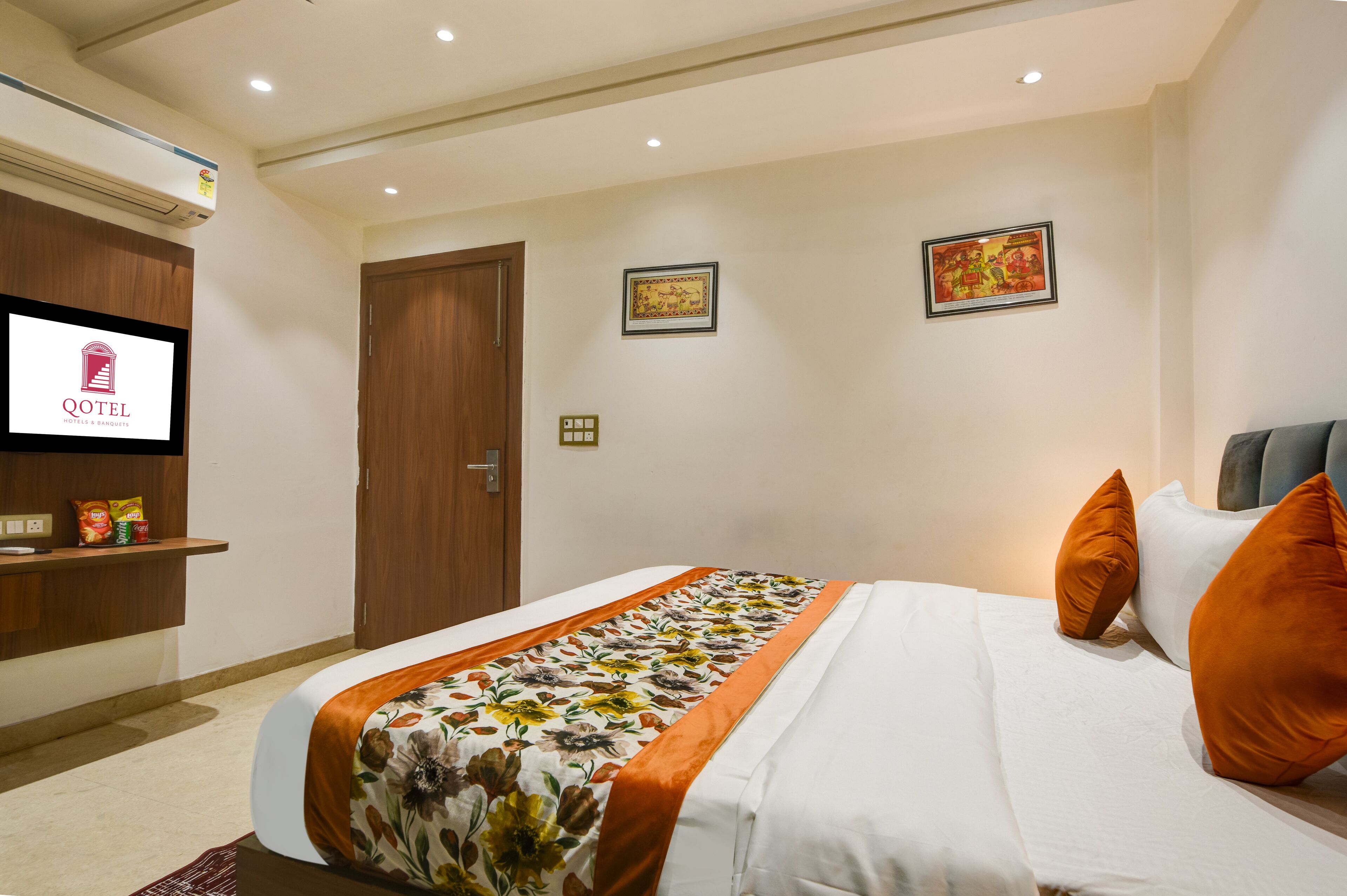 Habitación de hotel con una cama, un televisor, una puerta de madera y dos cuadros en la pared.