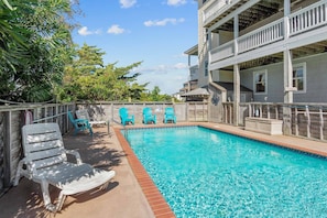 A swimming pool with blue water, surrounded by a wooden deck and a two-story building with balconies.
