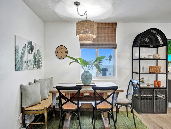 A dining area with a wooden table, black chairs, a woven chandelier, a clock, and a view of the beach through a window. - Oceanfront Condo on the Shores of Palm Beaches - Coastal Dawn (Palm Beach Shores)