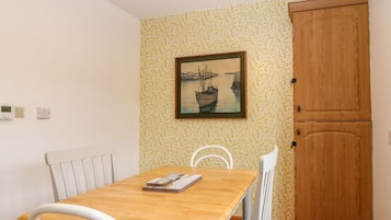 Un petit coin repas avec une table en bois et des chaises blanches, un tableau représentant des bateaux accroché au mur et une armoire en bois.