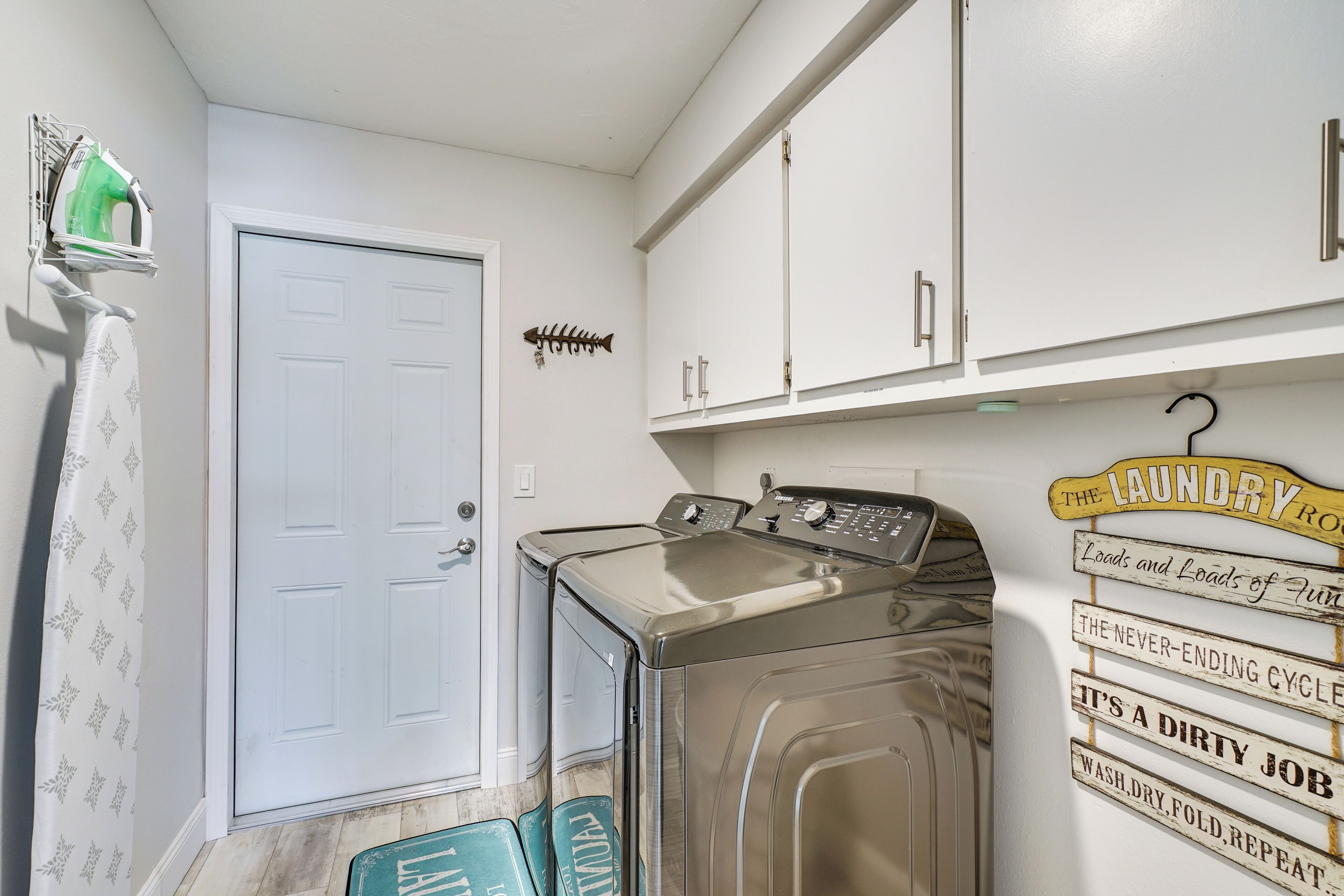 A laundry room with a washing machine, a dryer, and a wall-mounted ironing board. There are cabinets above the appliances and a door to the left.