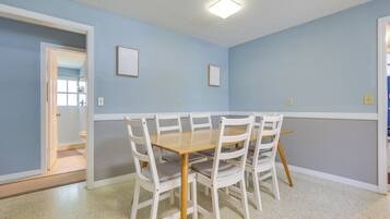 A dining area with a wooden table and white chairs, light blue walls, and a white door leading to a bathroom.