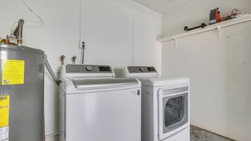 A laundry area with two washing machines and a dryer, a white wall, and a shelf with various items.