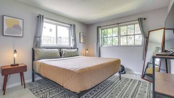 A bedroom with a bed, two windows with curtains, a bedside table with a lamp, a mirror, and a patterned rug.