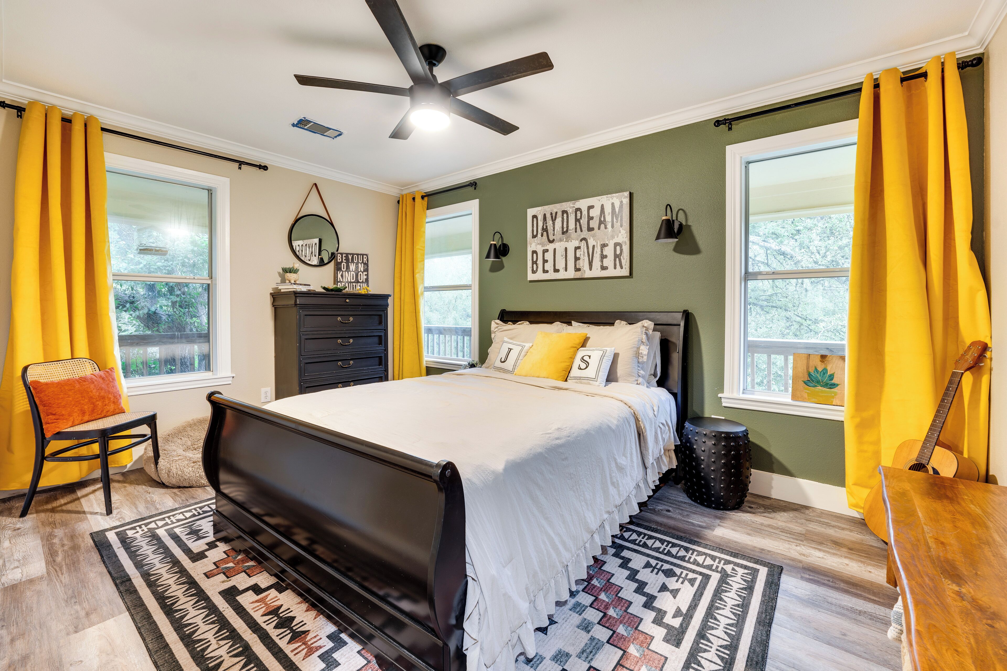 A bedroom with a bed, a ceiling fan, a dresser, a chair, a guitar, and a window with curtains.