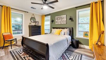 A bedroom with a bed, a ceiling fan, a dresser, a chair, a guitar, and a window with curtains.