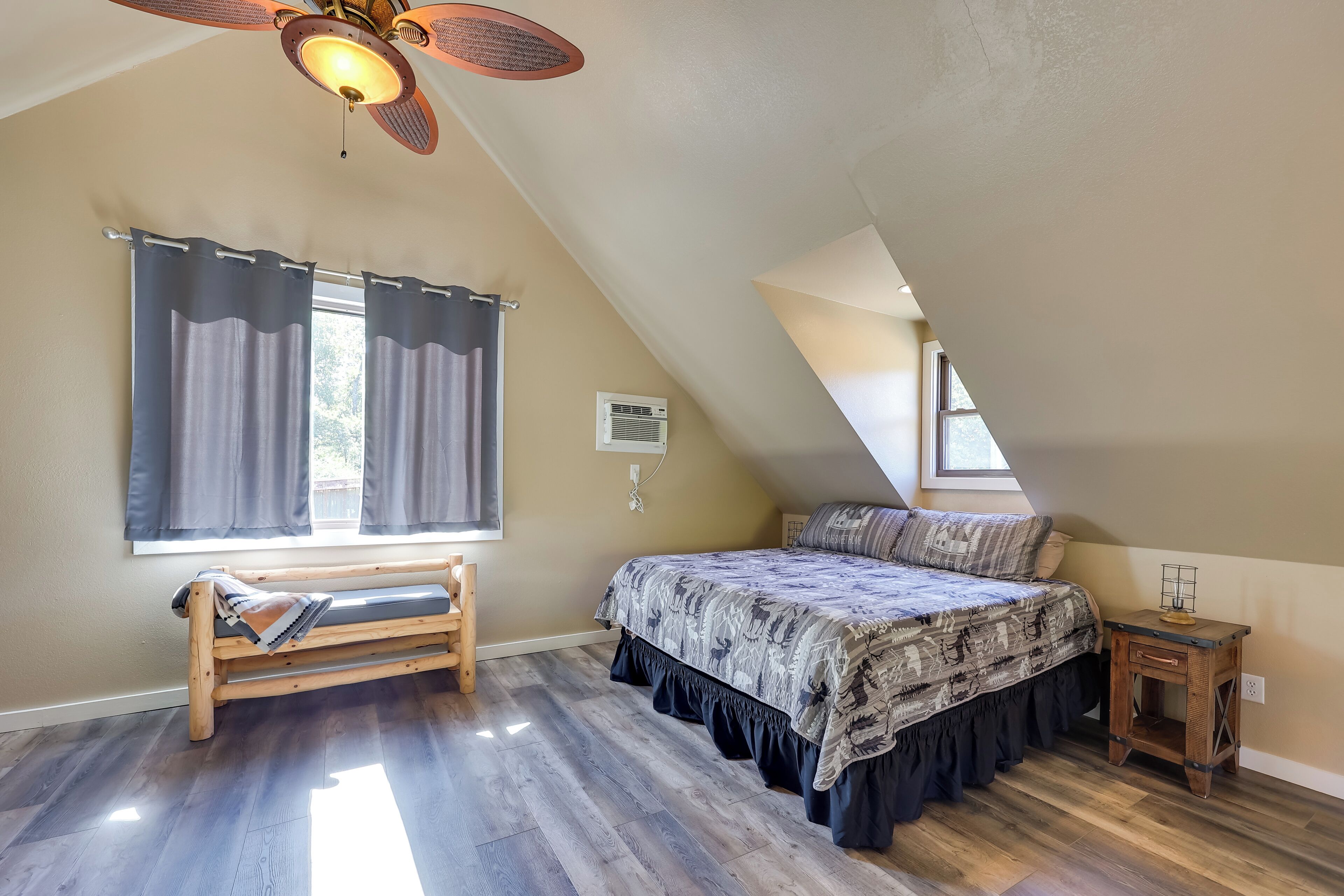 Une chambre avec un plafond en pente, un lit avec une literie à motifs, un banc, une petite table de chevet en bois et un ventilateur de plafond.