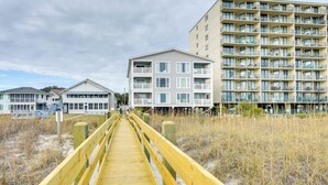 A wooden boardwalk leading to a beachfront building with multiple balconies and a second-floor balcony area.