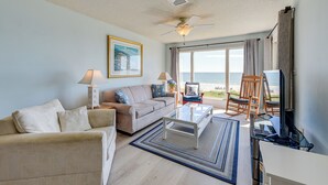 A living room with a sofa, coffee table, and a television. There is a window with a view of the beach.