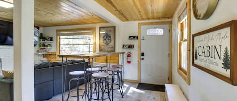 A cabin interior with a wooden ceiling, a dining area with stools, a sofa, and a wall-mounted clock.