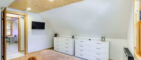 Ein Schlafzimmer mit schrägen Wänden, Holzdielenboden, einem großen Bett, zwei weißen Kommoden und einem an der Wand befestigten Fernseher.