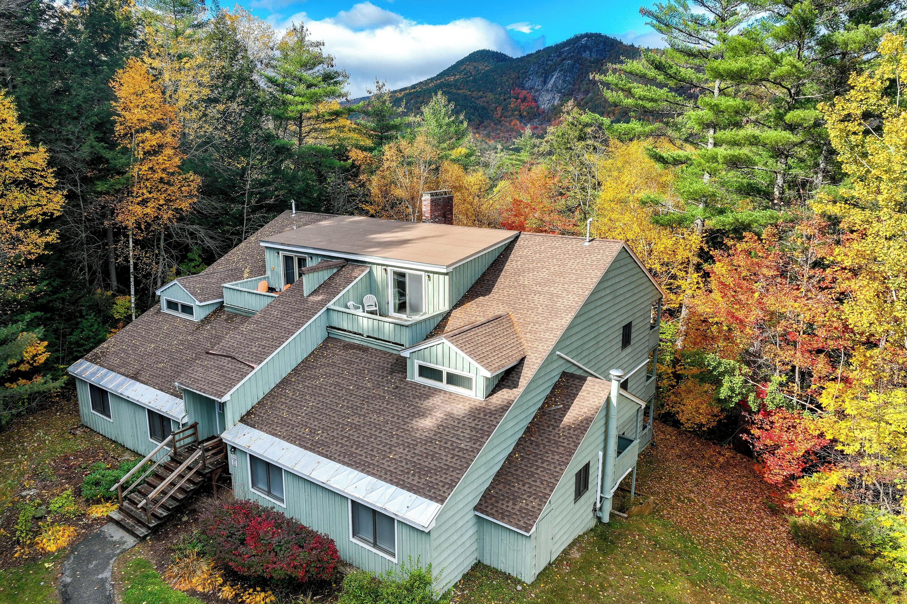 A two-story cabin with a steep roof, surrounded by autumn foliage and a mountainous backdrop.