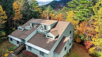 A two-story cabin with a steep roof, surrounded by autumn foliage and a mountainous backdrop.