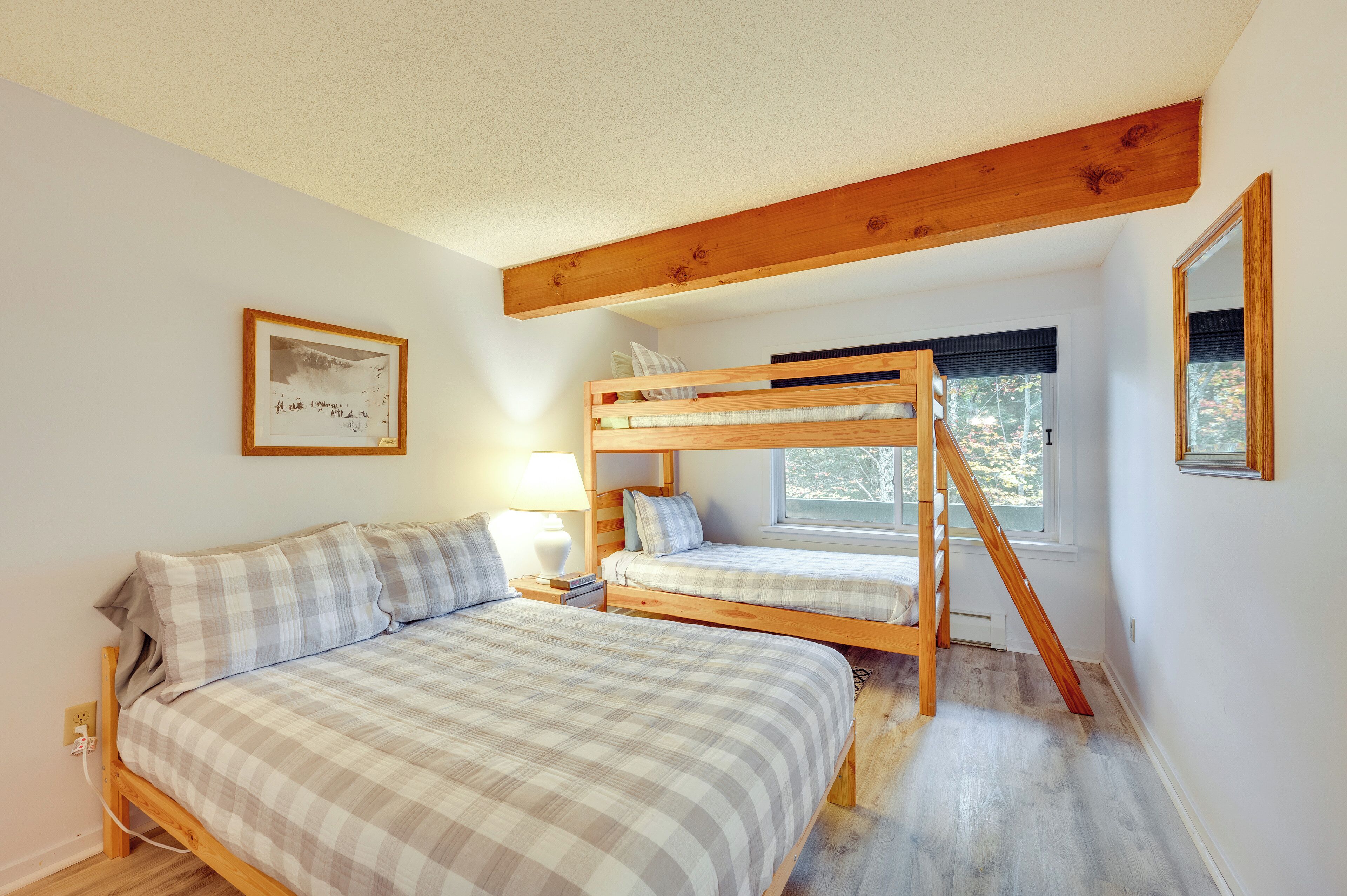 A bedroom with a bunk bed, a single bed, a bedside table with a lamp, a framed picture on the wall, and a window with a view of trees.